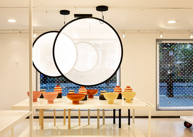 Two large circular pendant lamps with white diffusers and black frames hang above a display table with colorful ceramic vessels.