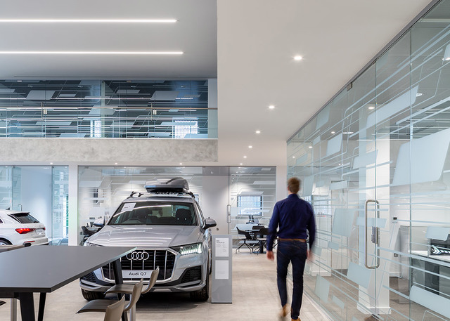 Audi showroom with recessed and linear ceiling lights illuminating a Q7 SUV display area, glass partitions, and open workspace.