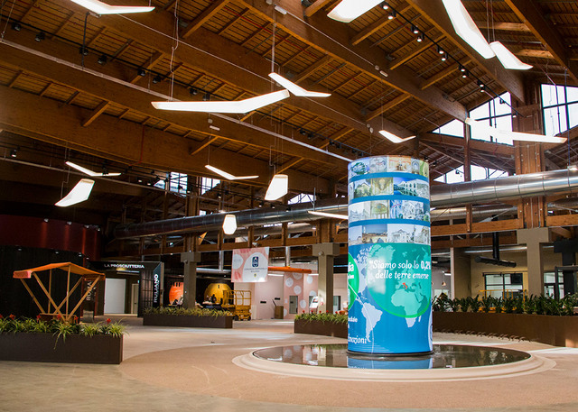 Large exhibition hall with wooden ceiling featuring Artemide pendant lights and a central cylindrical display with a world map graphic.
