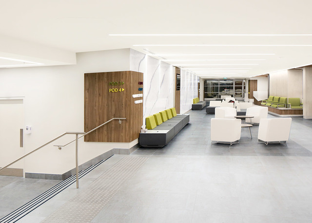 Modern hospital lobby with recessed linear ceiling lights, white seating, wood-paneled walls, and yellow-green accent cushions.