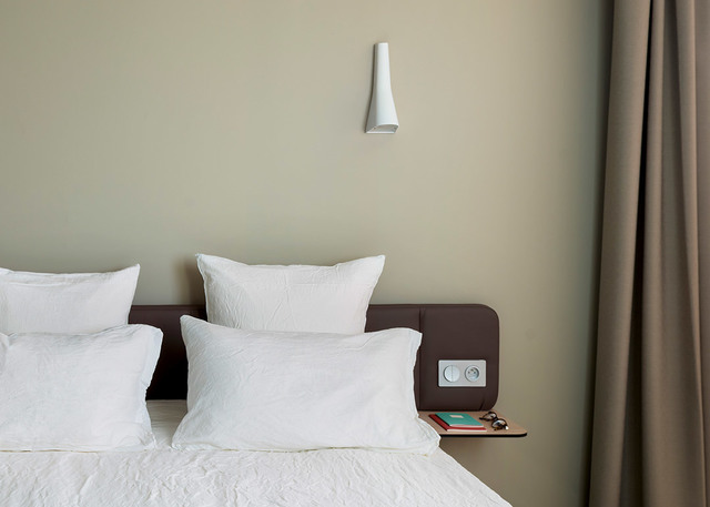 Artemide white conical wall lamp mounted above a bed headboard in a minimalist bedroom with sage green walls.