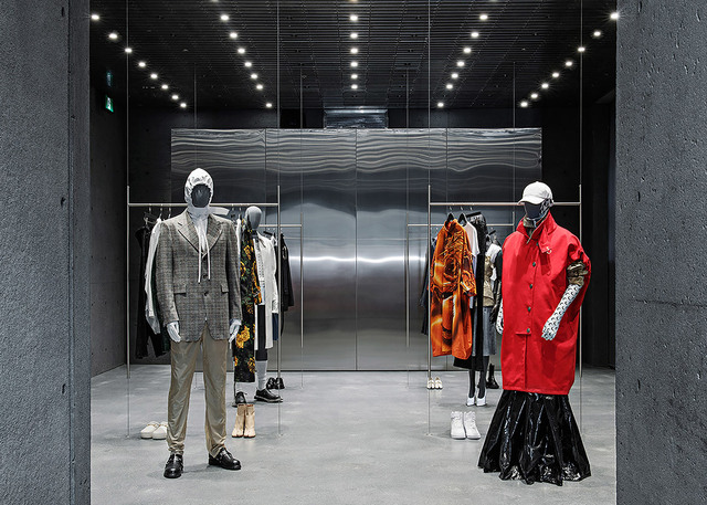 Fashion boutique interior with recessed ceiling spotlights illuminating two mannequins in designer outfits against metallic wall panels.
