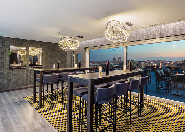 Rooftop bar with two Artemide circular LED pendant lights illuminating high tables and bar stools, with a panoramic city view at dusk.