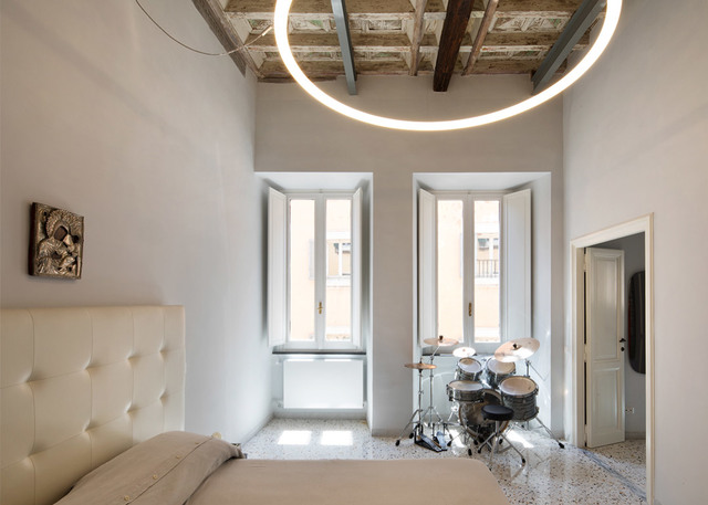Artemide circular LED pendant light illuminating a bright bedroom with exposed wooden beams, white walls, and a drum kit near tall windows.