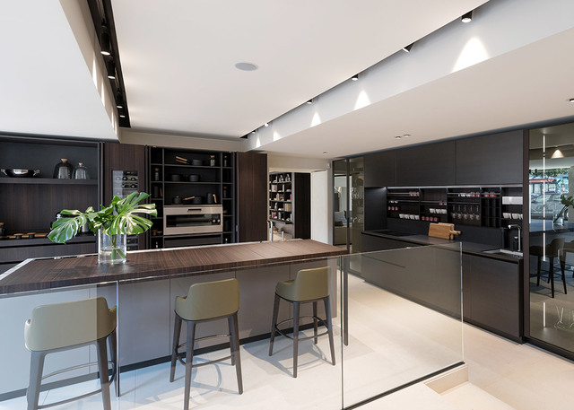 Modern kitchen with dark cabinetry lit by Artemide recessed spotlights on ceiling track, illuminating island with wood countertop and green stools