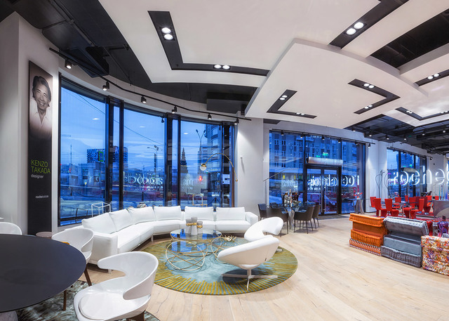 Modern hotel lobby with recessed ceiling spotlights, curved white sofa, designer chairs, and floor-to-ceiling windows overlooking a city street at dusk.