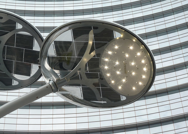 Two Artemide outdoor LED street lamps with circular heads on a steel pole, mounted against a curved glass building facade.