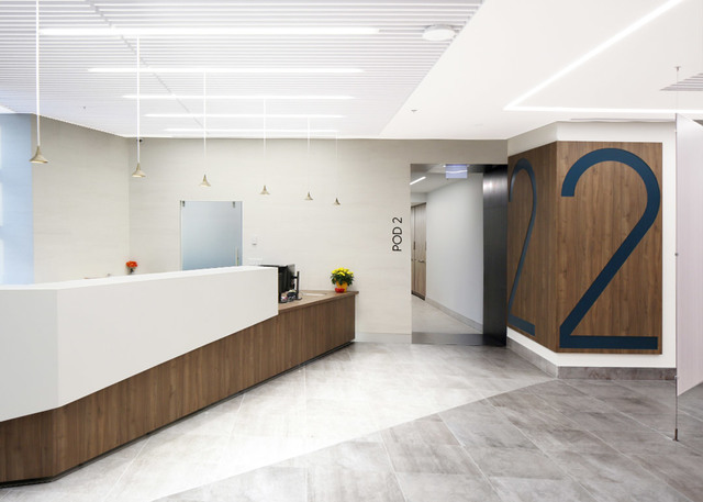 Modern healthcare reception area with white curved desk, wood accents, linear ceiling lights, and large number 2 sign marking Pod 2.