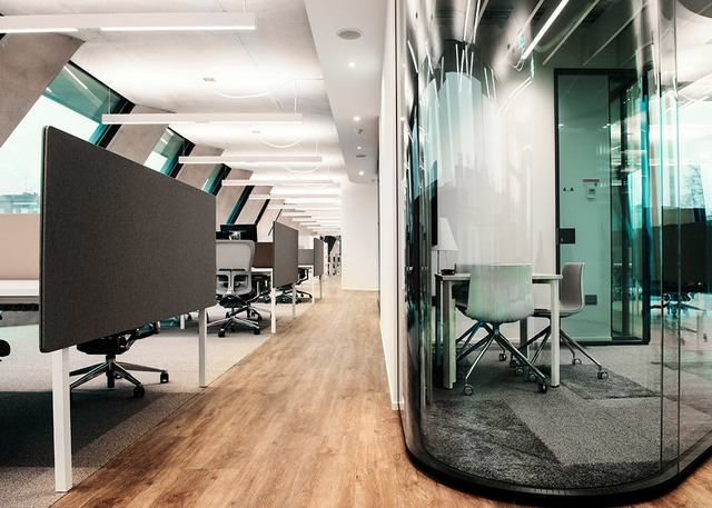 Modern open-plan office with workstations featuring acoustic panels, recessed ceiling lights, and a glass meeting pod.