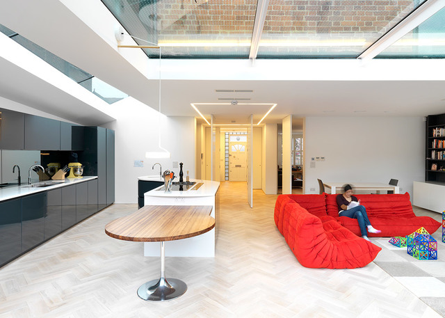 Modern open-plan living space with skylight, recessed linear LED ceiling lights, kitchen island, herringbone floor, and red sofa seating area.