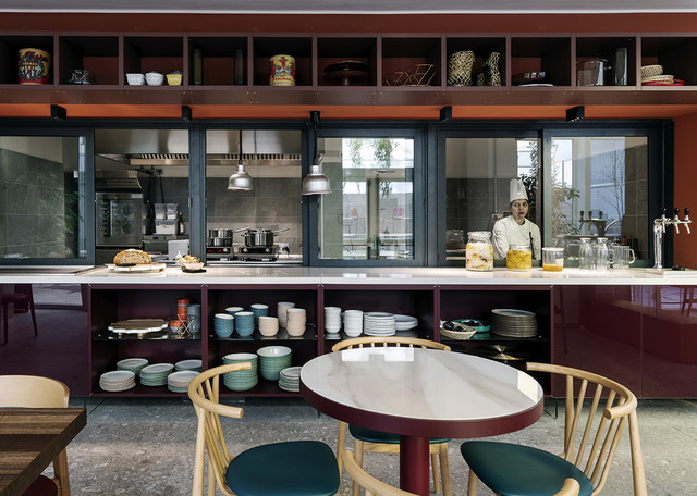 Modern restaurant interior with open kitchen, dark pendant lights, burgundy cabinetry displaying ceramics, and a round table with wooden chairs.