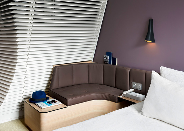 Hotel bedroom with Artemide black conical wall lamp above a curved brown leather sofa beside a white bed and venetian blinds
