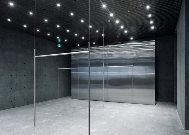 Modern interior with dark concrete walls, recessed ceiling spotlights, glass partitions, and a textured stainless steel wall panel.