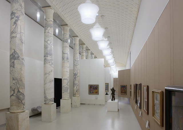 Museum gallery with marble columns and a row of spherical white ceiling lights illuminating artworks and sculptures along the walls.
