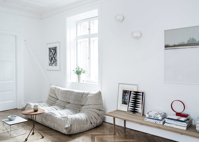Minimalist white living room with a grey sofa, wooden floor, bench with books, and a small round Artemide wall light.