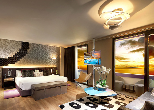 Luxury hotel bedroom featuring an Artemide ceiling lamp with swirling white rings, warm lighting, large window with sunset view.