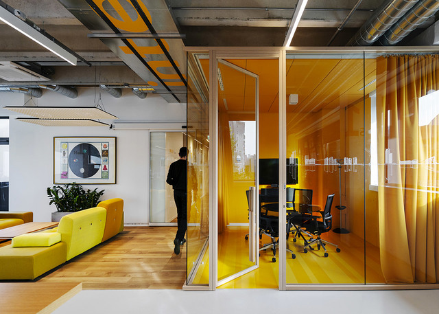 Modern office with yellow glass-walled meeting room, linear ceiling lights, and open lounge area with yellow modular sofa.