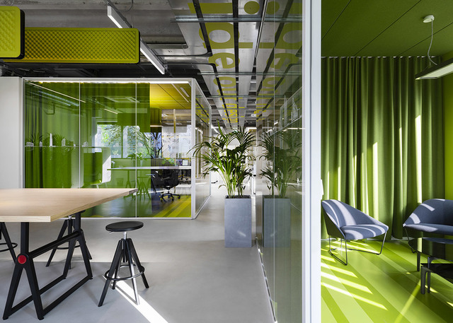 Modern open office with vibrant green accents, glass partitions, pendant lighting on exposed ceiling, workstations, and lush indoor plants