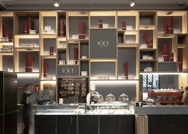Voce café interior with illuminated wooden shelving wall, track lighting above display cases, and staff serving pastries.