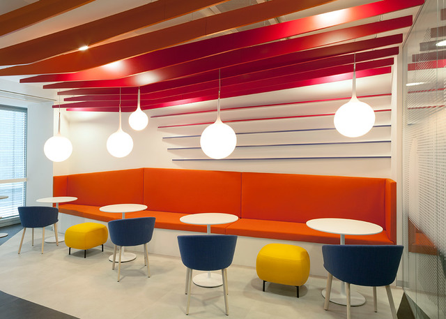 Modern lounge with five white spherical pendant lights over an orange sofa, colorful chairs and yellow ottomans beneath a red slatted ceiling