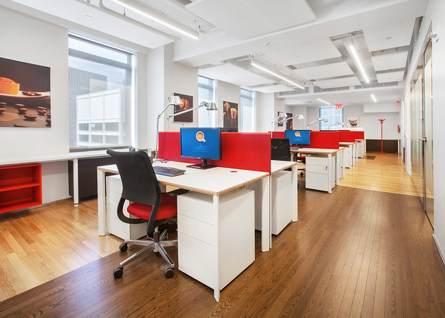 Modern open-plan office with white workstations, red dividers, task lamps, and linear ceiling lights illuminating a wood-floor workspace.