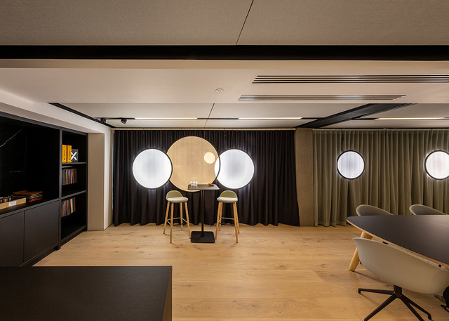 Modern office lounge with large circular glowing white pendant lights against dark curtains, wood floor, bar table and stools.