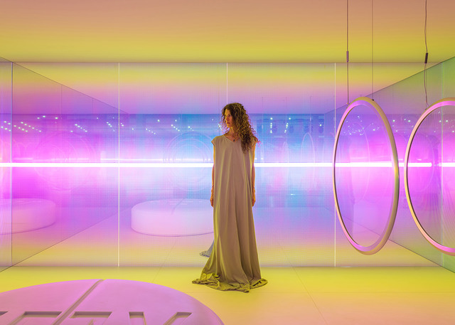 Woman in a mirrored room with vibrant pink and yellow gradient lighting, circular pendant lamps, and glowing neon light strips.