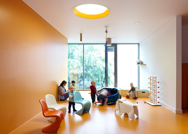 Bright children's playroom with a yellow-rimmed circular ceiling light, colorful chairs, and kids playing near large windows.