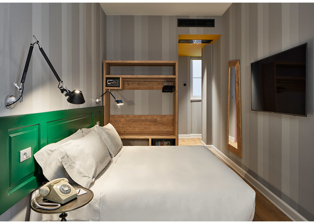 Hotel bedroom with Artemide Tolomeo wall lamp on grey striped wall, green headboard, white bedding, wooden shelving unit, and wall-mounted TV.