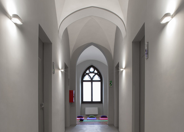 Gothic-arched corridor with white walls lit by Artemide wall sconces providing upward and downward light, leading to a pointed arch window.