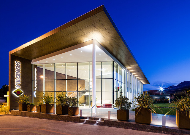 Modern commercial building exterior at dusk, featuring large glass facade, cantilevered roof with warm recessed lighting, and landscaped entrance.
