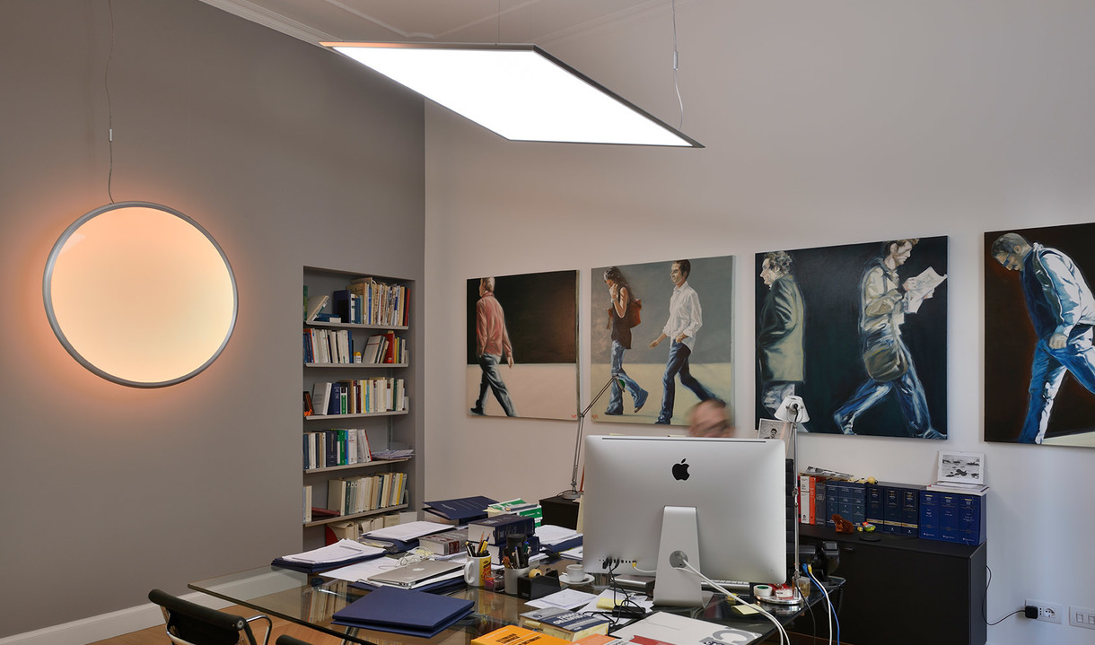 Office workspace with two Artemide lights: a round wall lamp glowing warmly and a rectangular LED pendant above a desk with computer and artwork.