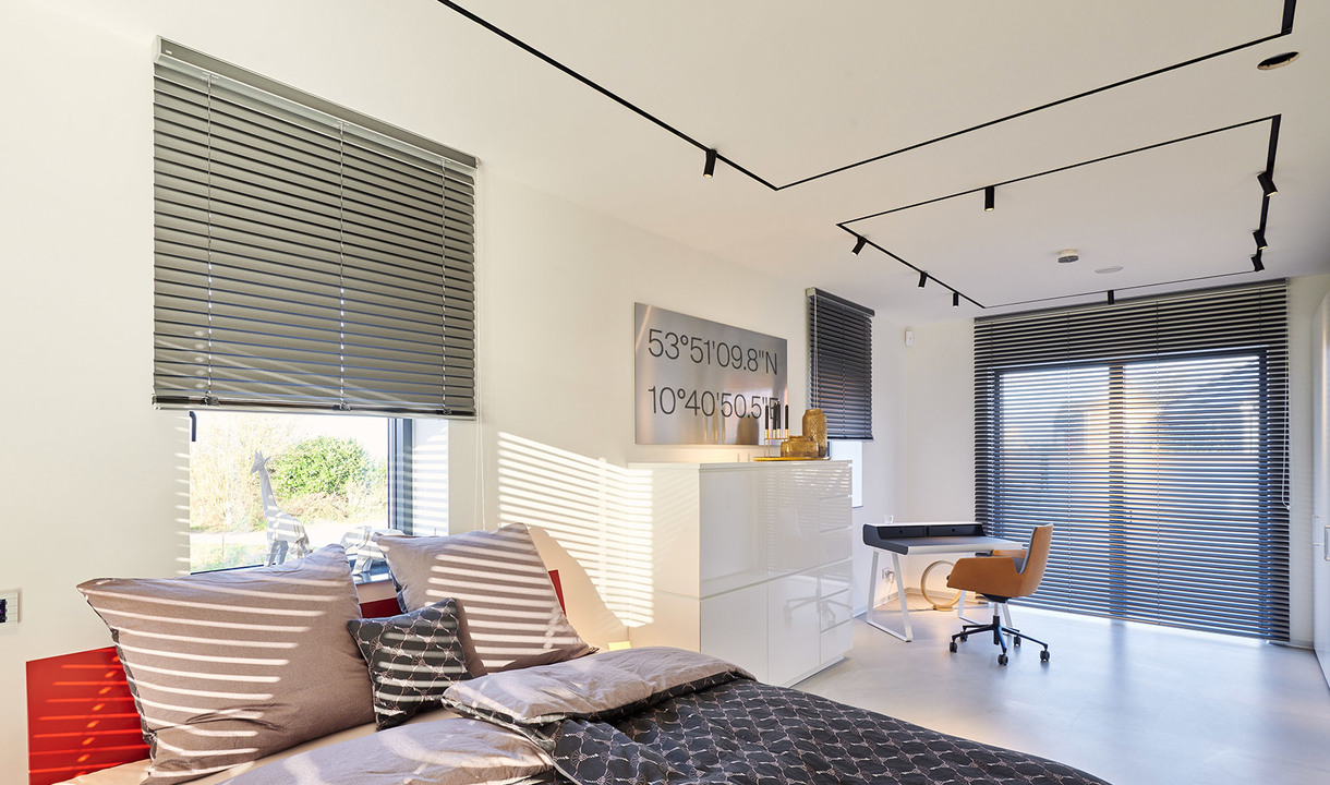 Modern bedroom with Artemide track lighting on ceiling, gray venetian blinds, bed with cushions, and a desk workspace area