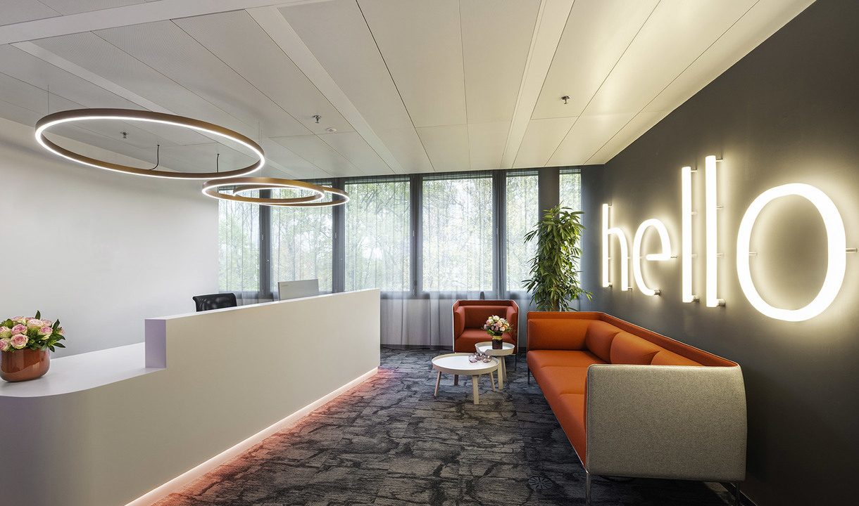 Modern office reception with Artemide circular pendant lights, orange sofa, white desk, and illuminated 'hello' sign on dark wall
