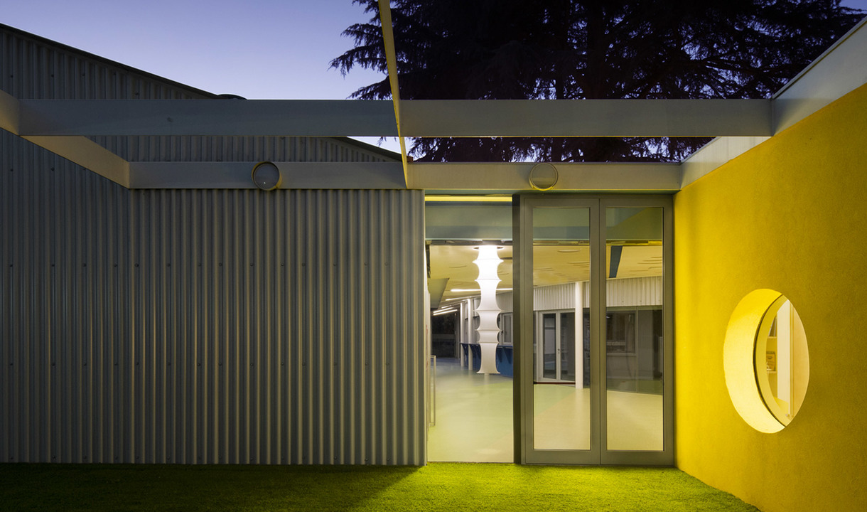 Modern building exterior at dusk with yellow accent wall, circular window, and glowing white columnar lamp visible through glass doors.