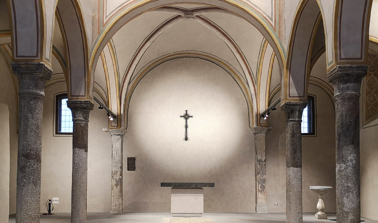 Historic chapel interior with vaulted ceilings, stone columns, and a crucifix on the apse wall, lit by discreet spotlights