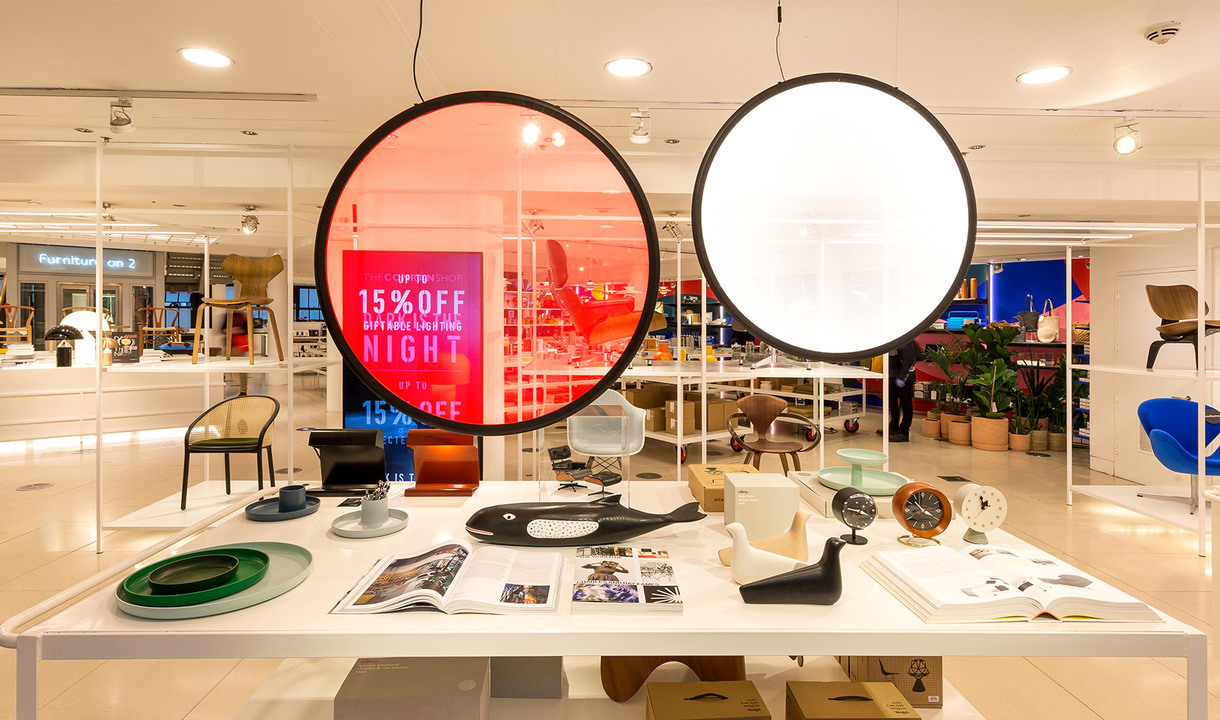 Design store interior with two large circular pendant lamps, one glowing red, one white, above a display table with design objects.