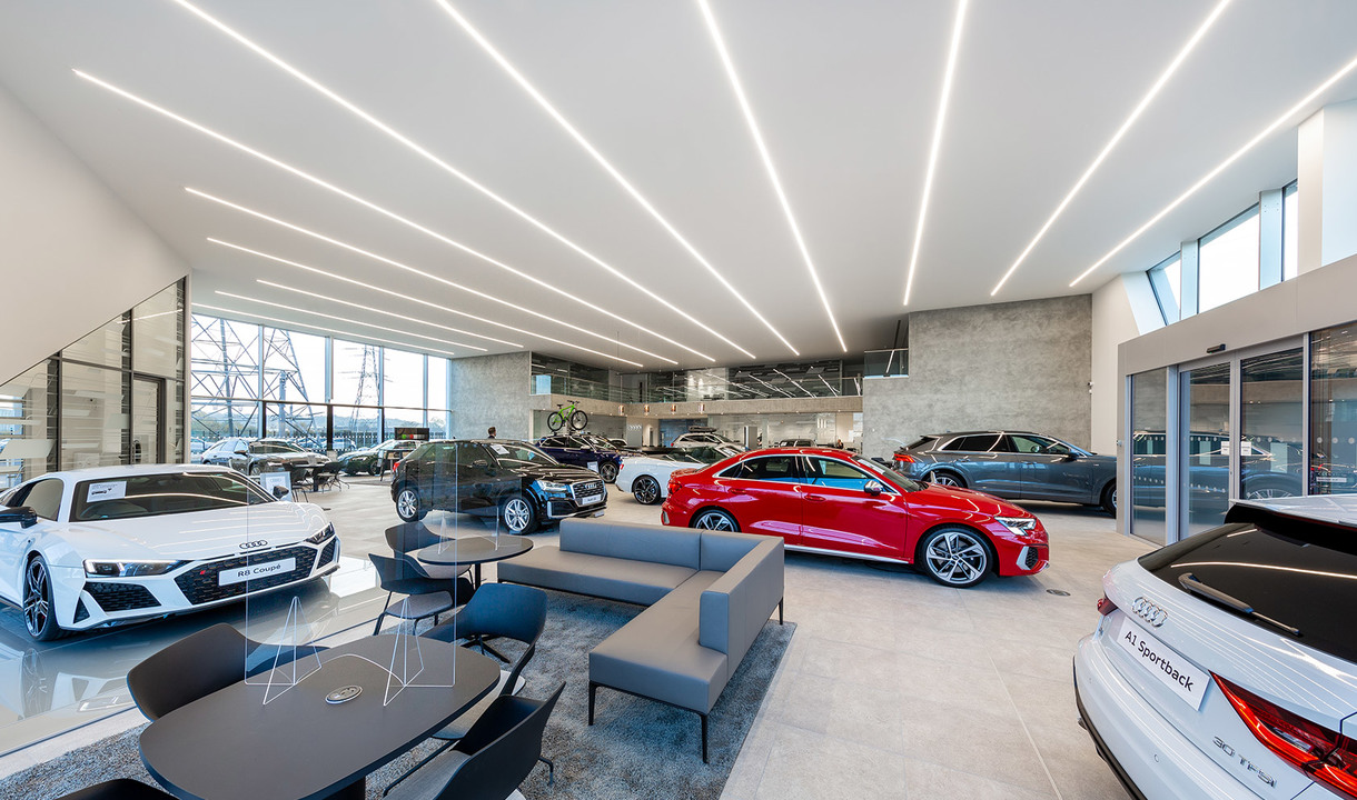 Audi car showroom with linear LED ceiling lights illuminating displayed vehicles on a polished floor with lounge seating area