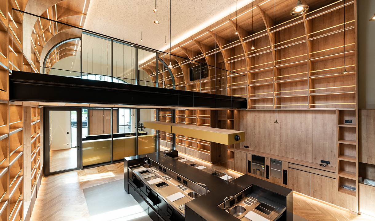 Double-height library interior with warm wood shelving, herringbone floor, pendant lights, and a dark kitchen island below a mezzanine.