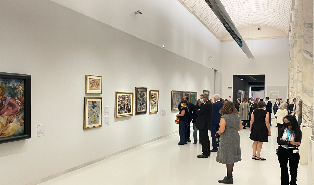 Museum gallery with visitors viewing colorful paintings on white walls, lit by recessed ceiling spotlights illuminating the artwork.