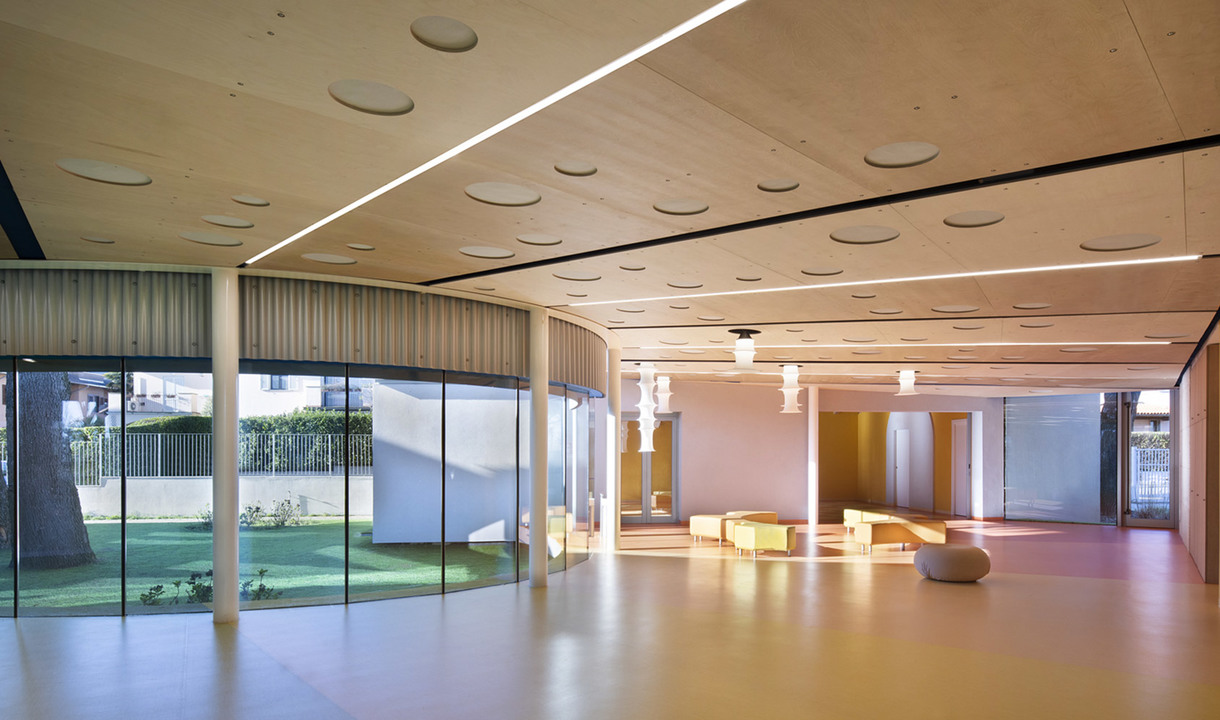 Spacious modern interior with warm wood ceiling, recessed linear LED lighting, pendant lamps, and floor-to-ceiling glass walls overlooking a garden