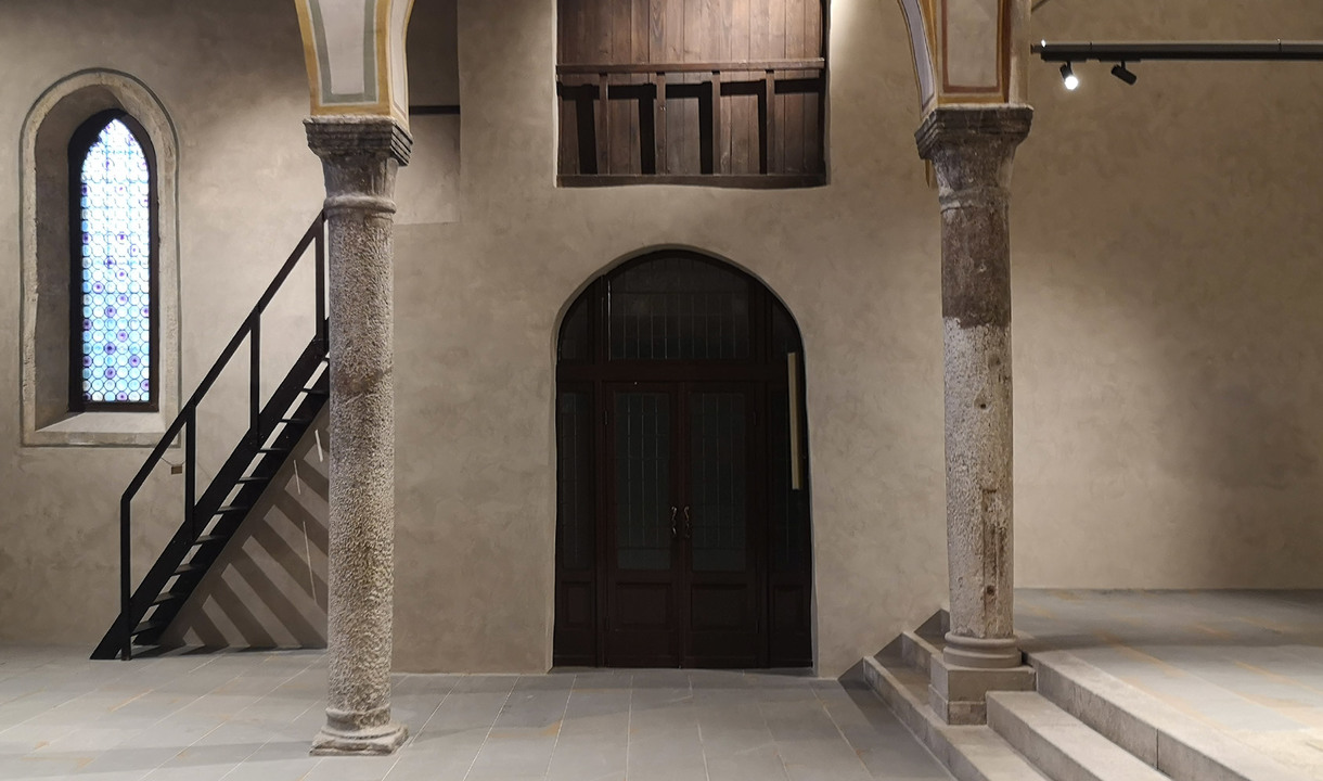 Historic interior with stone columns, arched wooden door, stained glass window, black staircase, and Artemide track spotlights on ceiling.