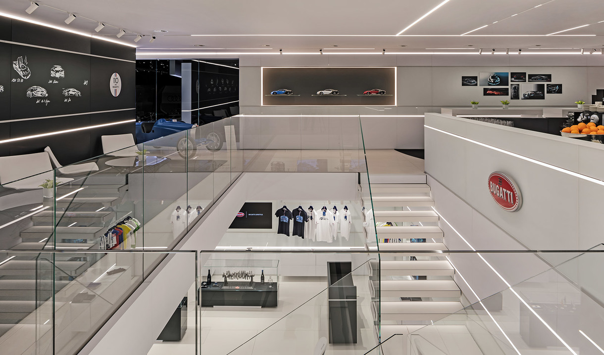 Bugatti showroom interior with two levels, white minimalist design, linear LED lighting, glass railings, and luxury merchandise displays