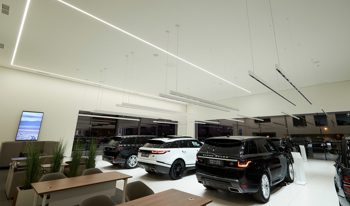 Car showroom with luxury SUVs illuminated by Artemide linear pendant and ceiling LED light fixtures creating bright, even lighting.