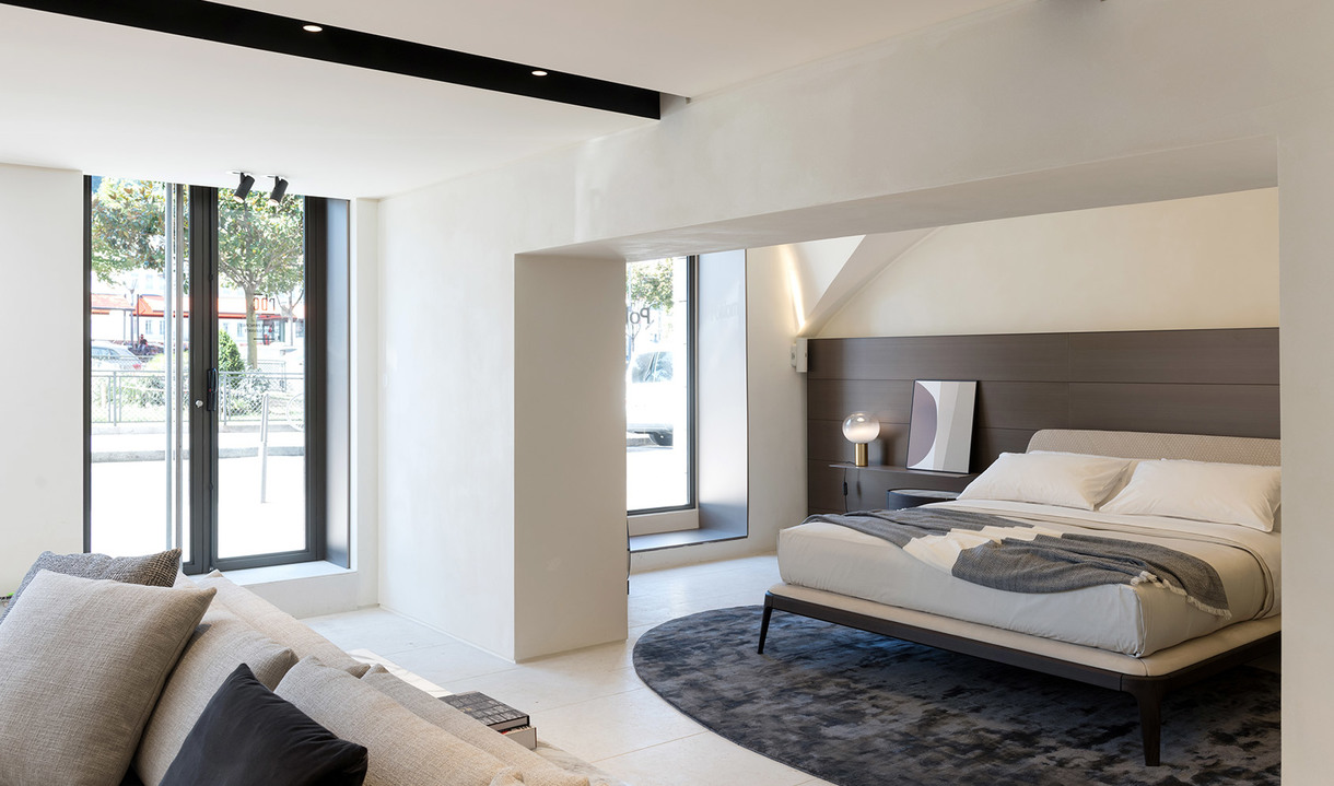 Modern bedroom with minimalist design, featuring a table lamp on a nightstand, white walls, dark headboard, and circular dark rug