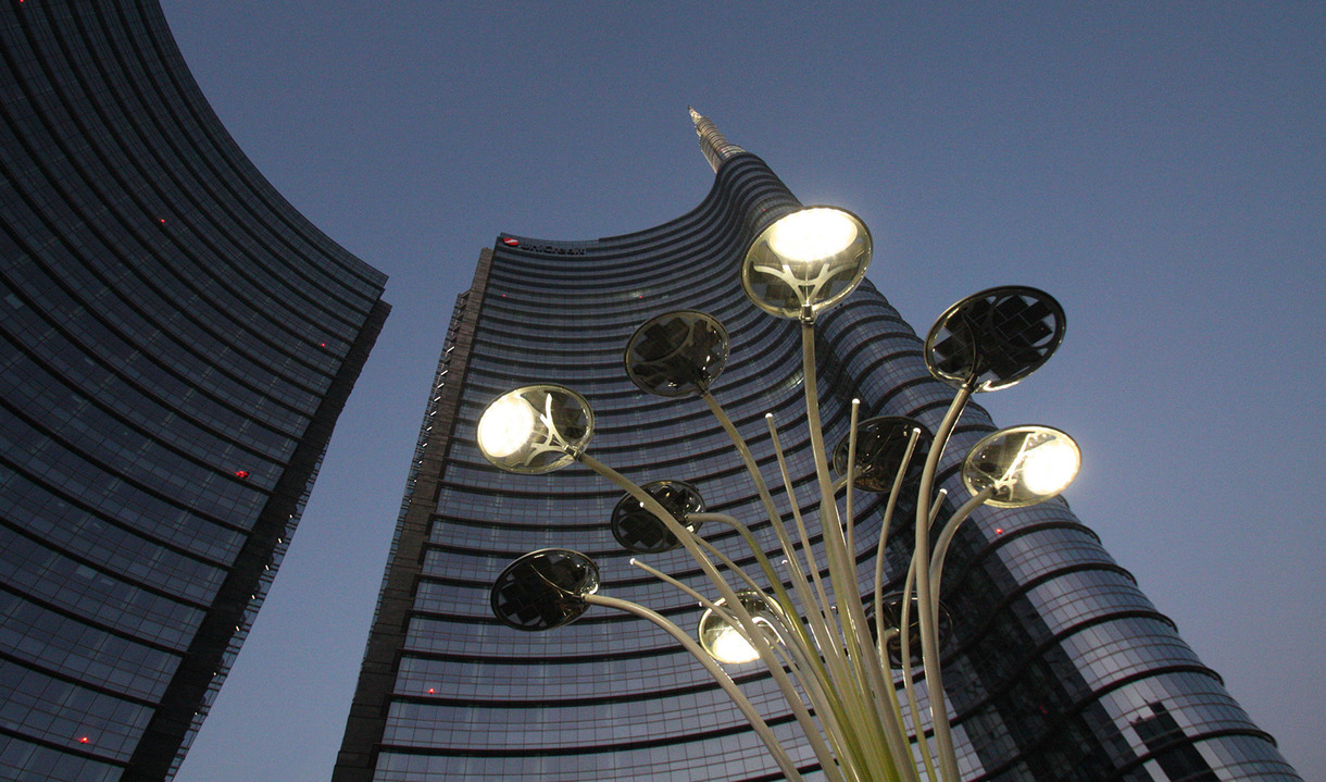 Artemide outdoor sculptural lamp installation with multiple stems and flower-shaped heads, illuminated at dusk in front of a curved glass skyscraper in Milan.