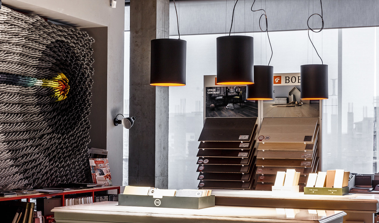 Three black cylindrical pendant lamps with gold inner shades hanging in a modern showroom with concrete columns and display shelves.
