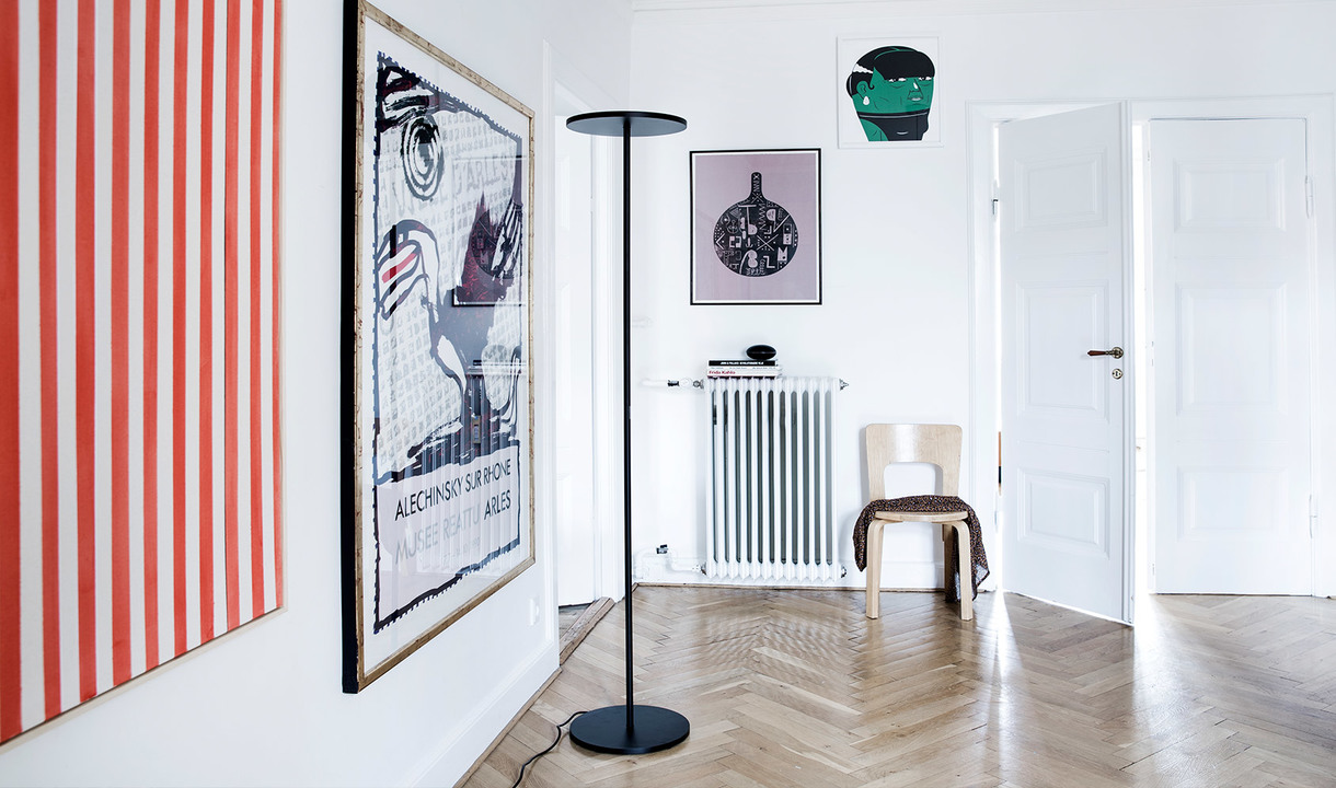 Black floor lamp with flat circular shade in a bright art-filled room with herringbone parquet floor and white walls.