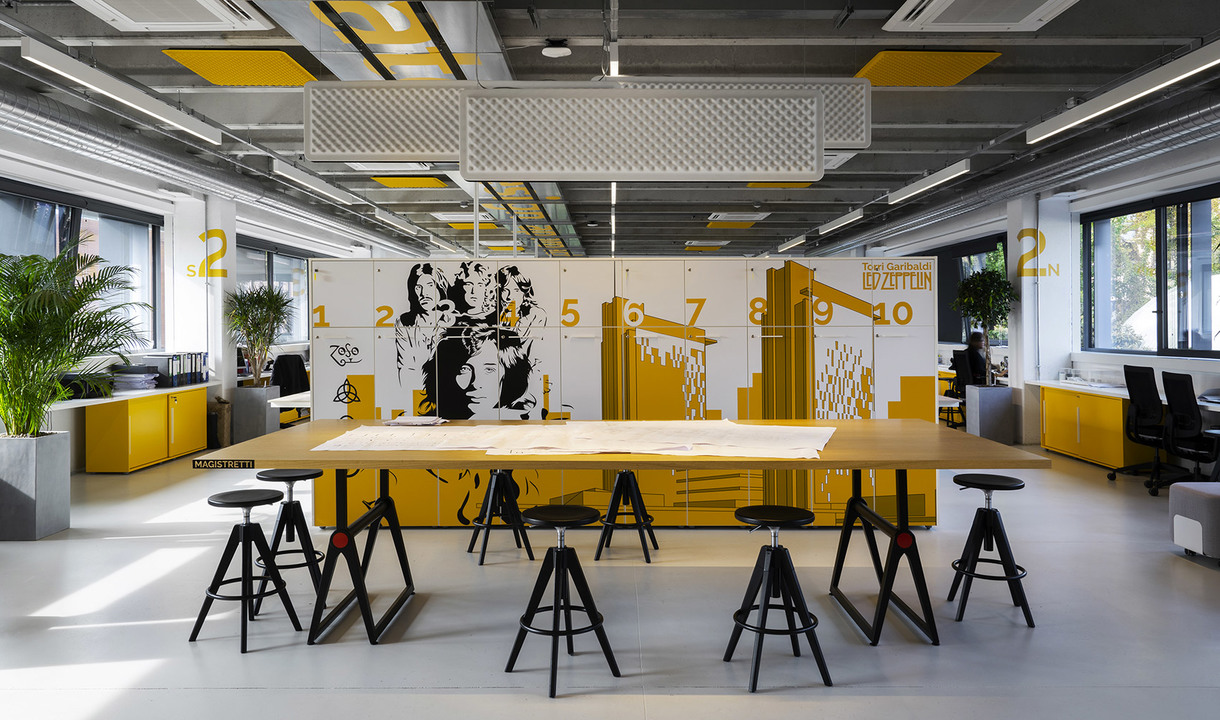 Modern office workspace with yellow accents, large worktable, black stools, rectangular pendant lights, and bold graphic mural wall.