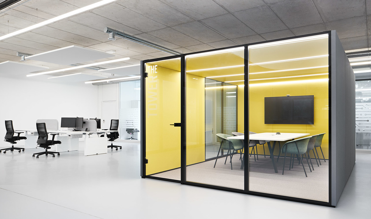 Modern office with open workstations and a glass-walled meeting pod featuring yellow walls, linear LED ceiling lights, and a conference table.